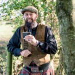 Schuggie at the side of a wood wearing a tweed cap and raising his hipflask. Slàinte Mhath, Schuggie, from Schuggies-Ceilidhs.